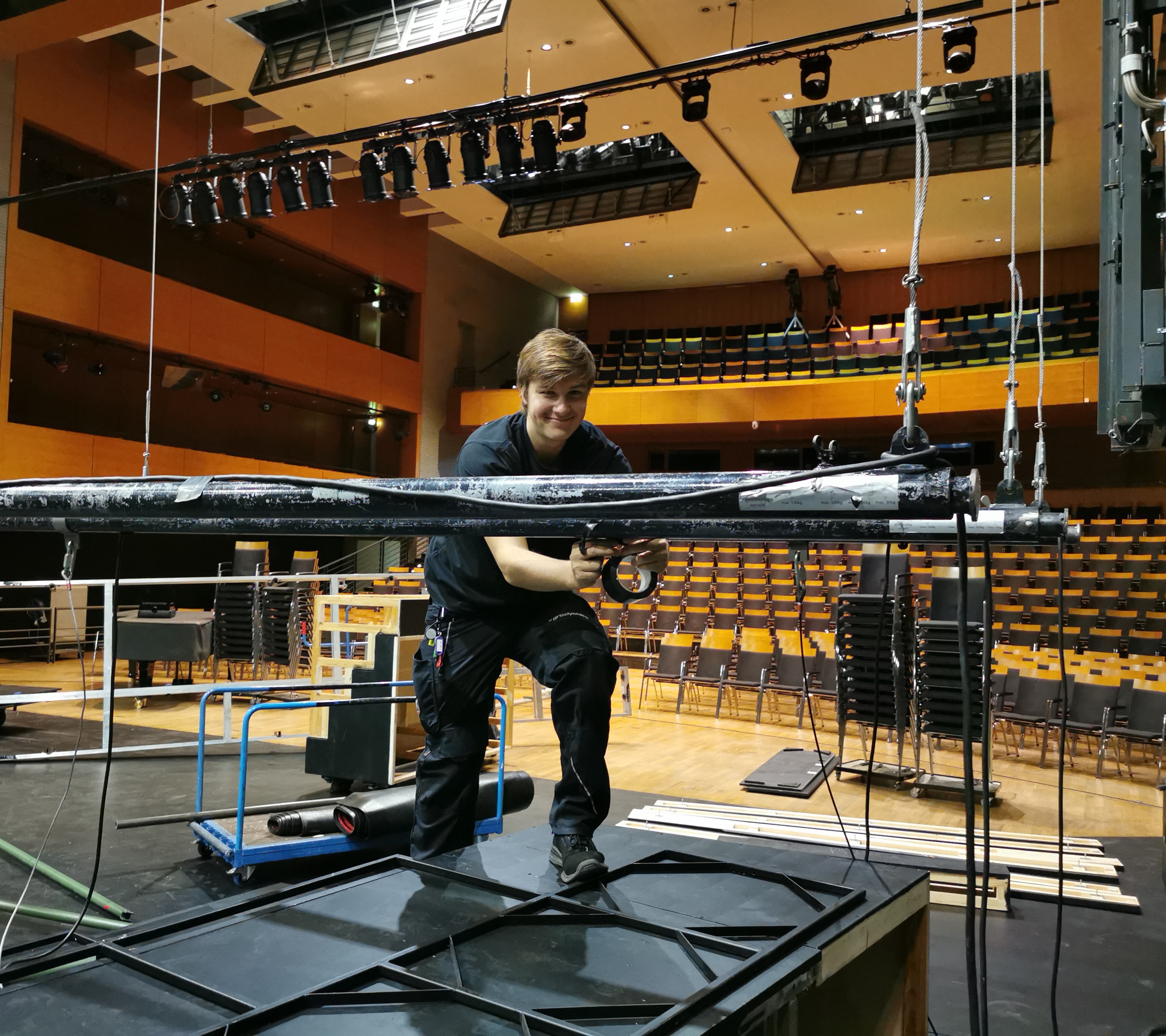 ein junger Techniker montiert etwas an Haltestangen, im Hintergund der llere Zuschauerraum